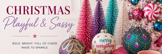 A festive Christmas holiday decor arrangement featuring colorful mini trees, patterned ornaments, ribbons, a pink mug reading 'Sleigh all day,' and decorative text on the left that says 'CHRISTMAS Playful & Sassy.' No person, animal, or real environment is present; this is a styled product scene, so subject_type is Product/Packaging rather than Lifestyle.