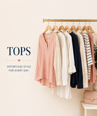 A set of women's tops in soft neutral tones hanging on wooden hangers from a gold clothing rack against a warm beige wall. The collection includes a dusty pink top, a cream lace blouse, a navy blouse, and striped tops.