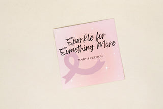 A square pink card or sticker with the text 'Sparkle for Something More' and 'MARY'S VERSION' above a pink ribbon graphic, placed on a plain light gray surface.